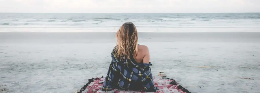 Frau sitzt mit dem Rücken zur Kamera am Strand und blickt aufs Meer